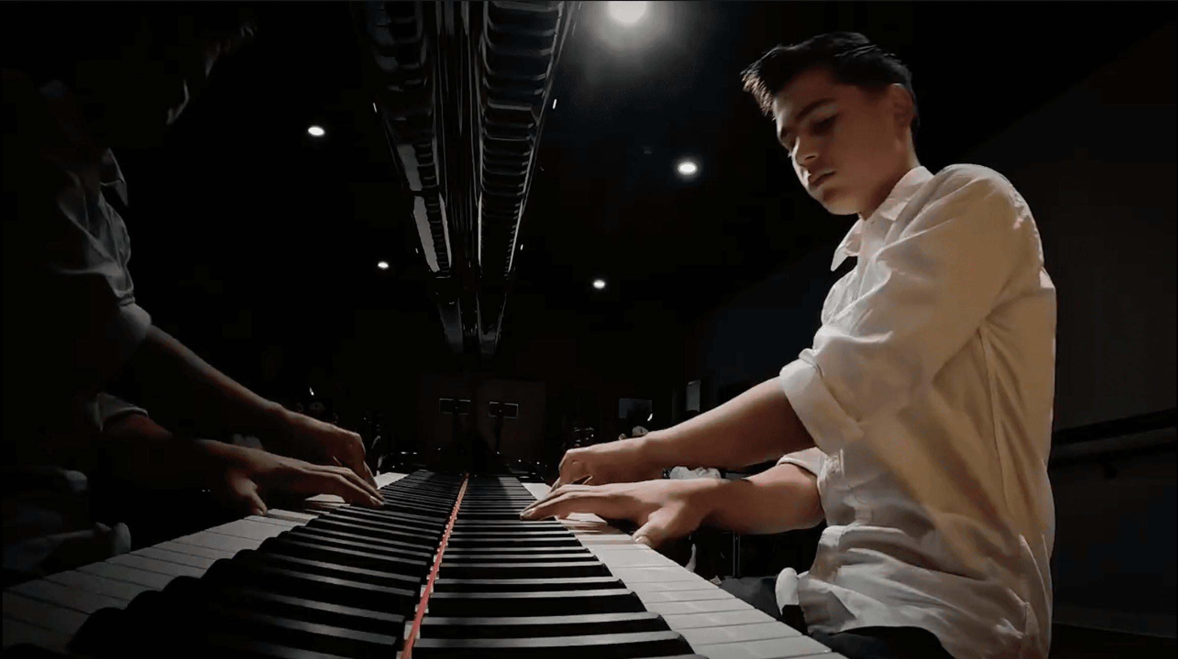 Focused young man playing a grand piano under spotlights, reflected in its polished surface.
